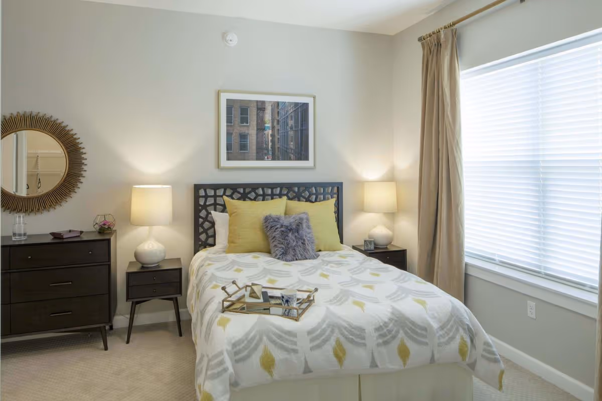 A neatly made bed with a patterned white and yellow comforter and three pillows, two yellow and one gray fluffy, in a bright bedroom. The bed has a decorative dark headboard and a tray with a cup and small items on it. On each side of the bed are dark wooden nightstands with white lamps. To the left is a matching dresser with a round decorative mirror above it. A large window with beige curtains is on the right wall, allowing natural light to fill the room.