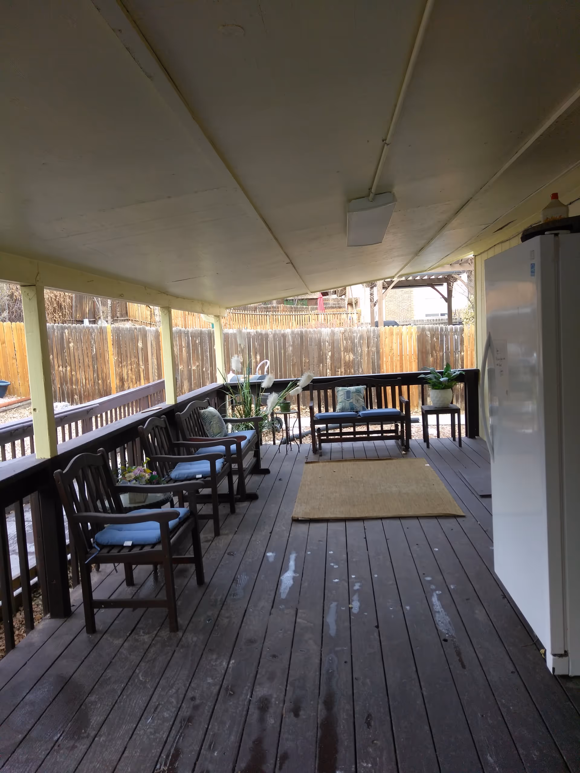 Covered outdoor wooden deck with several wooden chairs and benches with blue cushions, potted plants, a beige rug in the center, and a white refrigerator on the right side. The deck is enclosed by a wooden fence.