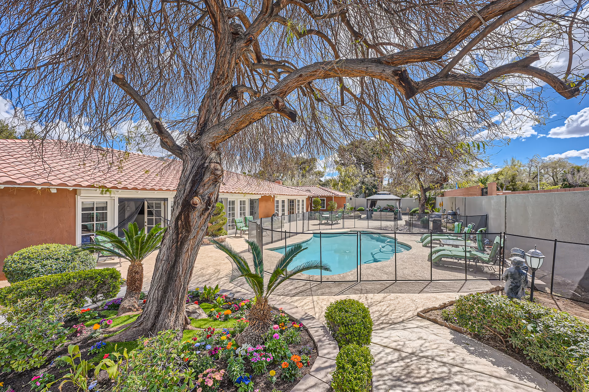 A landscaped courtyard featuring a fenced swimming pool, lounge chairs, a large tree, and colorful flower beds in front of a single-story building.