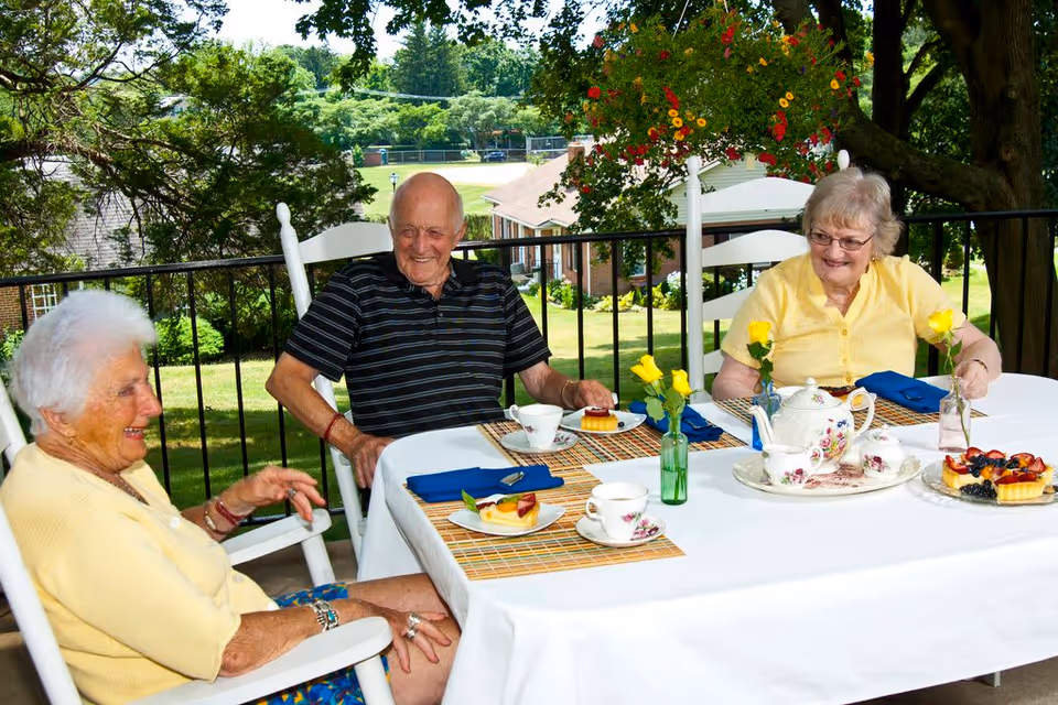 Three elderly people sitting around a table outdoors on a patio, enjoying tea and dessert. The table is covered with a white tablecloth and set with a floral teapot, teacups, small vases with yellow flowers, and plates with fruit-topped desserts. The background shows greenery, trees, and buildings under a clear sky.