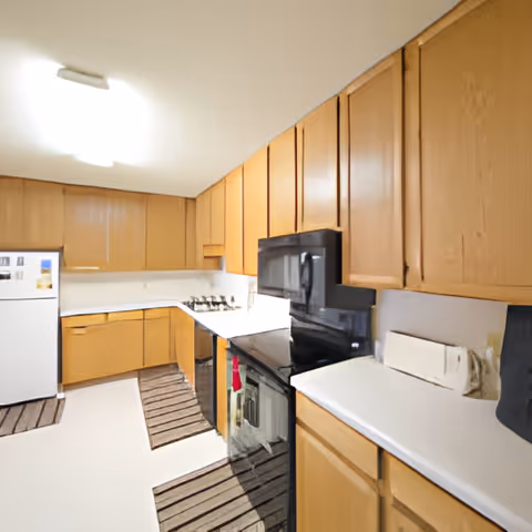 A kitchen with wooden cabinets, white countertops, a white refrigerator, a black microwave, and a black oven. There are striped rugs on the floor and a paper towel holder on the counter.
