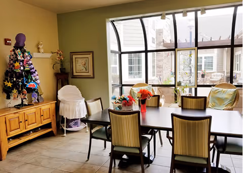 A bright dining room with a rectangular table surrounded by six chairs. The table has small flower arrangements in vases. There is a wooden sideboard with decorative items and a colorful quilt or fabric draped over a mannequin or stand. Large windows provide a view of an outdoor patio area with additional seating.