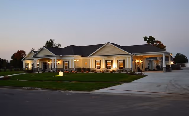 Exterior view of a single-story retirement home building at dusk with warm lights illuminating the front porch and windows. The building has a beige and stone facade with a dark roof, surrounded by a well-maintained lawn and a paved driveway.