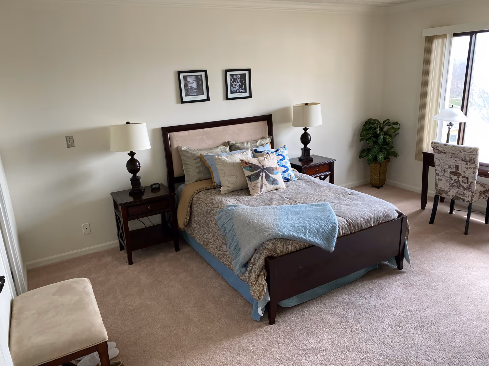 A neatly made bed with multiple pillows and a light blue throw blanket in a bedroom. There are two dark wooden nightstands with matching lamps on either side of the bed. Two framed pictures hang on the wall above the headboard. A small cushioned bench is positioned near the foot of the bed. A window with vertical blinds lets in natural light, and a desk with a patterned chair and a lamp is placed near the window. A potted plant is in the corner of the room.
