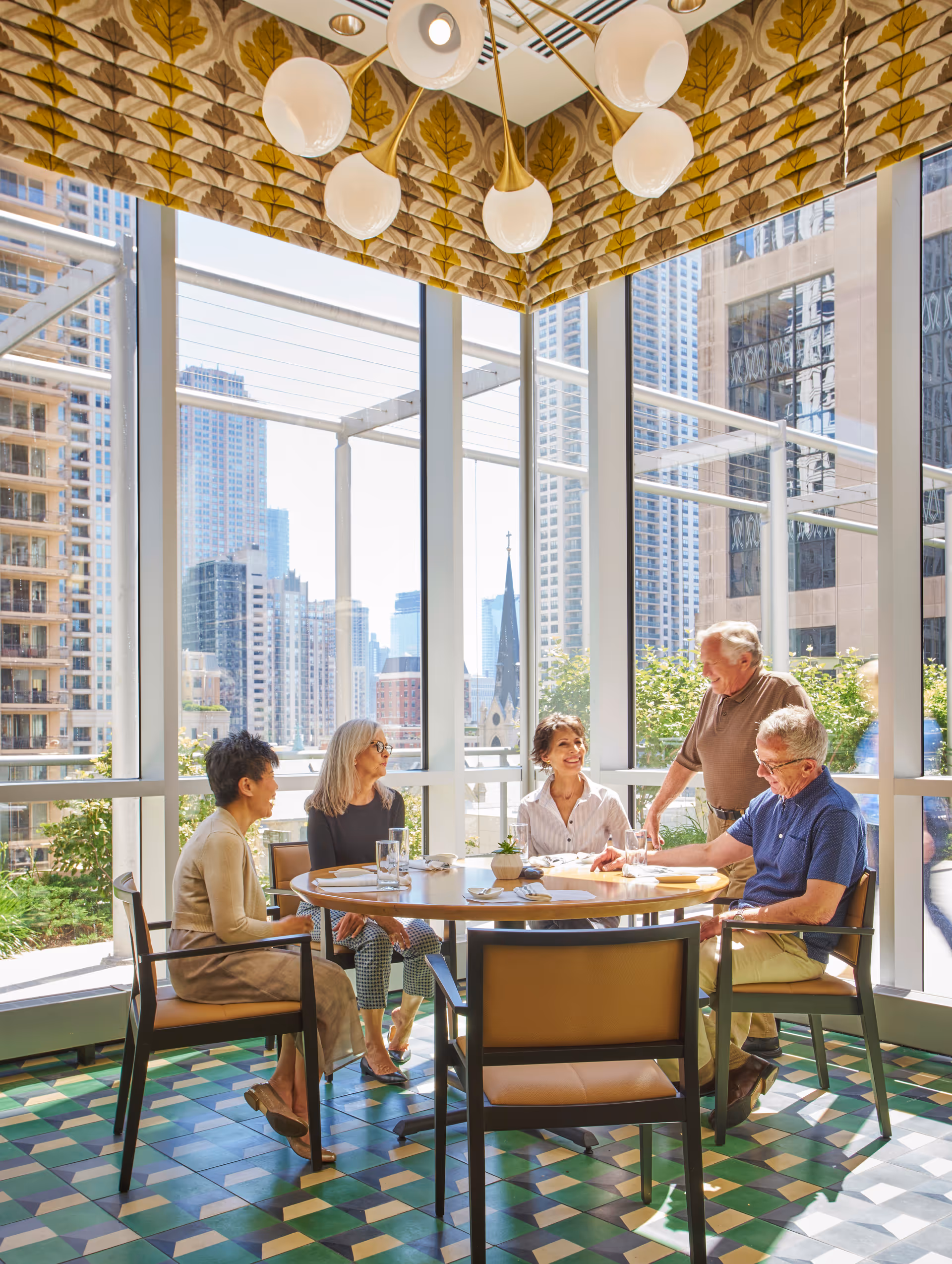 A group of five elderly people sitting and standing around a round dining table in a bright room with large floor-to-ceiling windows showing a cityscape. The room has patterned tile flooring and a decorative ceiling with a modern chandelier. The people are engaged in conversation and appear to be enjoying each other's company.