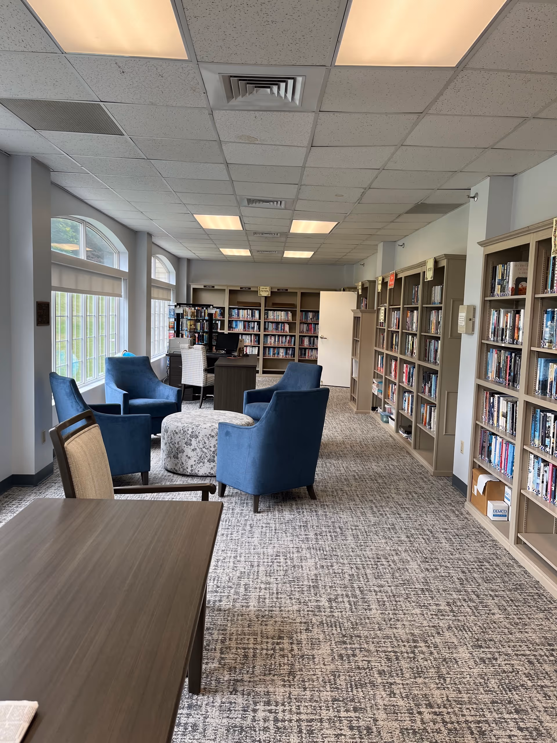 Interior view of a library room in Avery Heights with large windows on the left side letting in natural light. The room has several bookshelves filled with books along the right wall and at the far end. There are four blue armchairs arranged around a round, patterned ottoman in the center of the room. A desk with a computer and a chair is positioned near the bookshelves at the far end. The floor is carpeted with a patterned design, and the ceiling has recessed lighting panels.