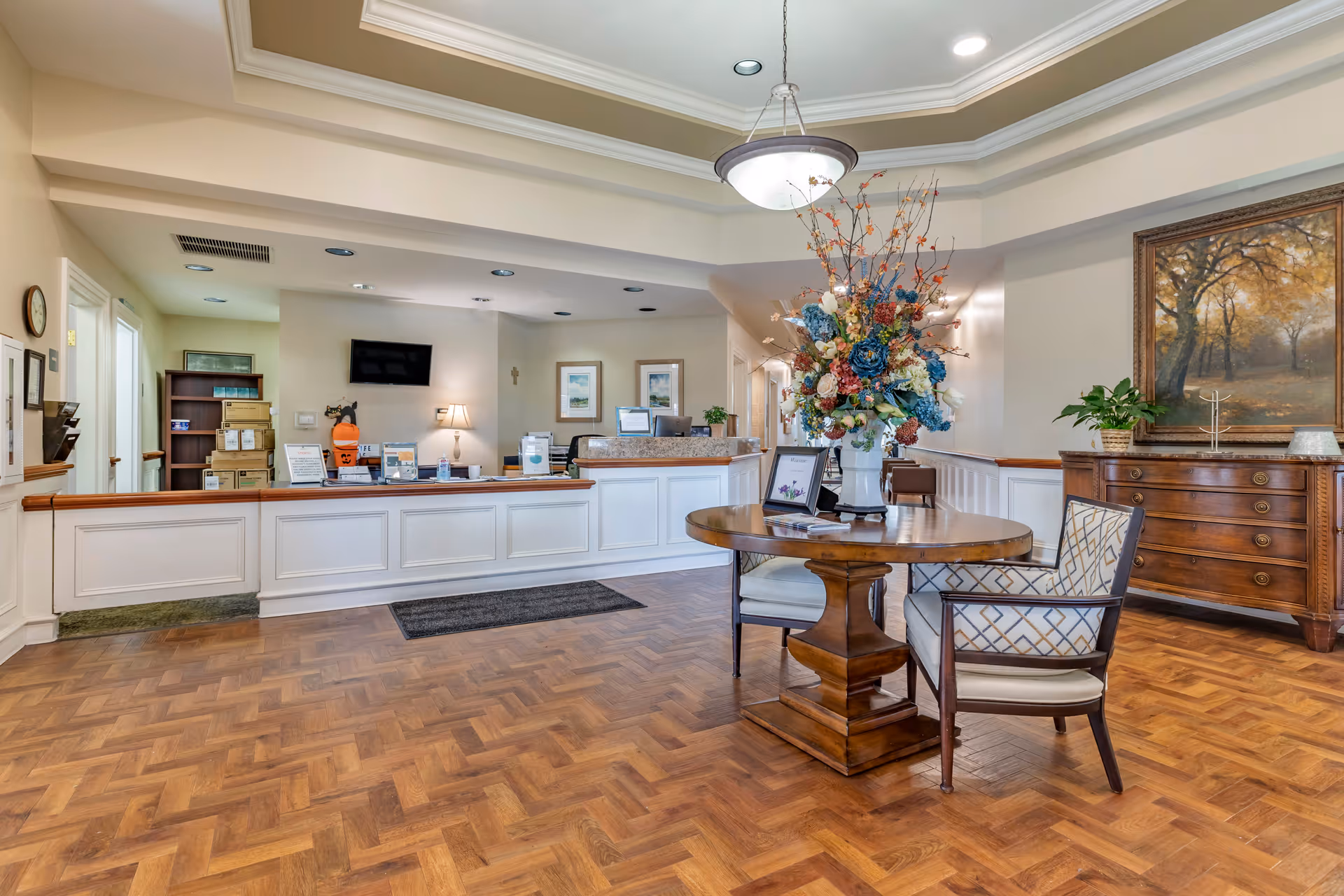 Reception area of a senior living facility with a wooden floor, a round wooden table with a large floral arrangement in the center, two upholstered chairs, a wooden dresser with a plant and a framed painting of a forest scene on the wall, and a long white reception desk with lamps, a TV mounted on the wall, and various informational materials.
