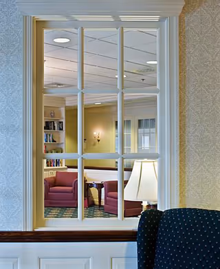 View through a multi-pane window into a cozy sitting area with two red armchairs, a small wooden table, a lamp, and bookshelves. The room has soft lighting and patterned carpet.