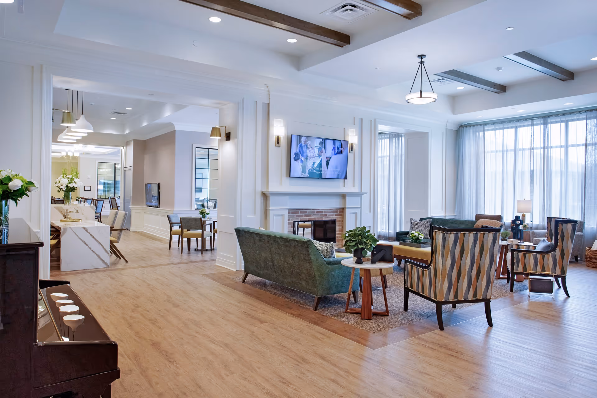 A bright and spacious senior living facility common area with wooden flooring, a green sofa, patterned armchairs, a fireplace with a mounted TV above it, and large windows with sheer curtains allowing natural light to fill the room. There is a piano on the left side and a dining area visible in the background with tables, chairs, and pendant lighting.