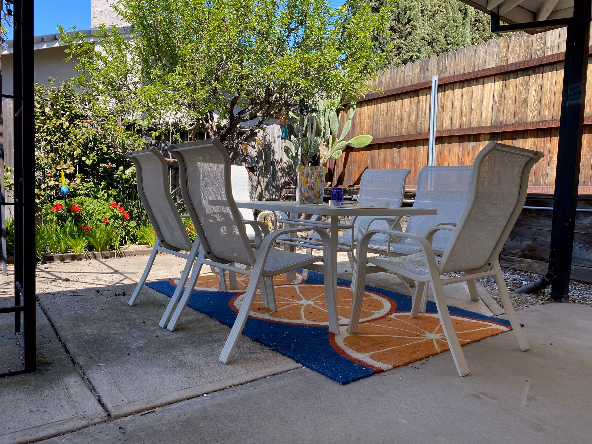 Outdoor patio area with a white table and six white chairs arranged around it on a colorful rug with orange slice designs. The patio is surrounded by a wooden fence, green plants, and a tree providing shade.