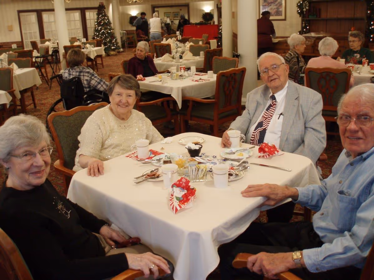 A group of elderly people sitting around a table in a dining room decorated for Christmas, with a Christmas tree and festive decorations in the background. The table is set with cups, plates, and holiday-themed napkins.