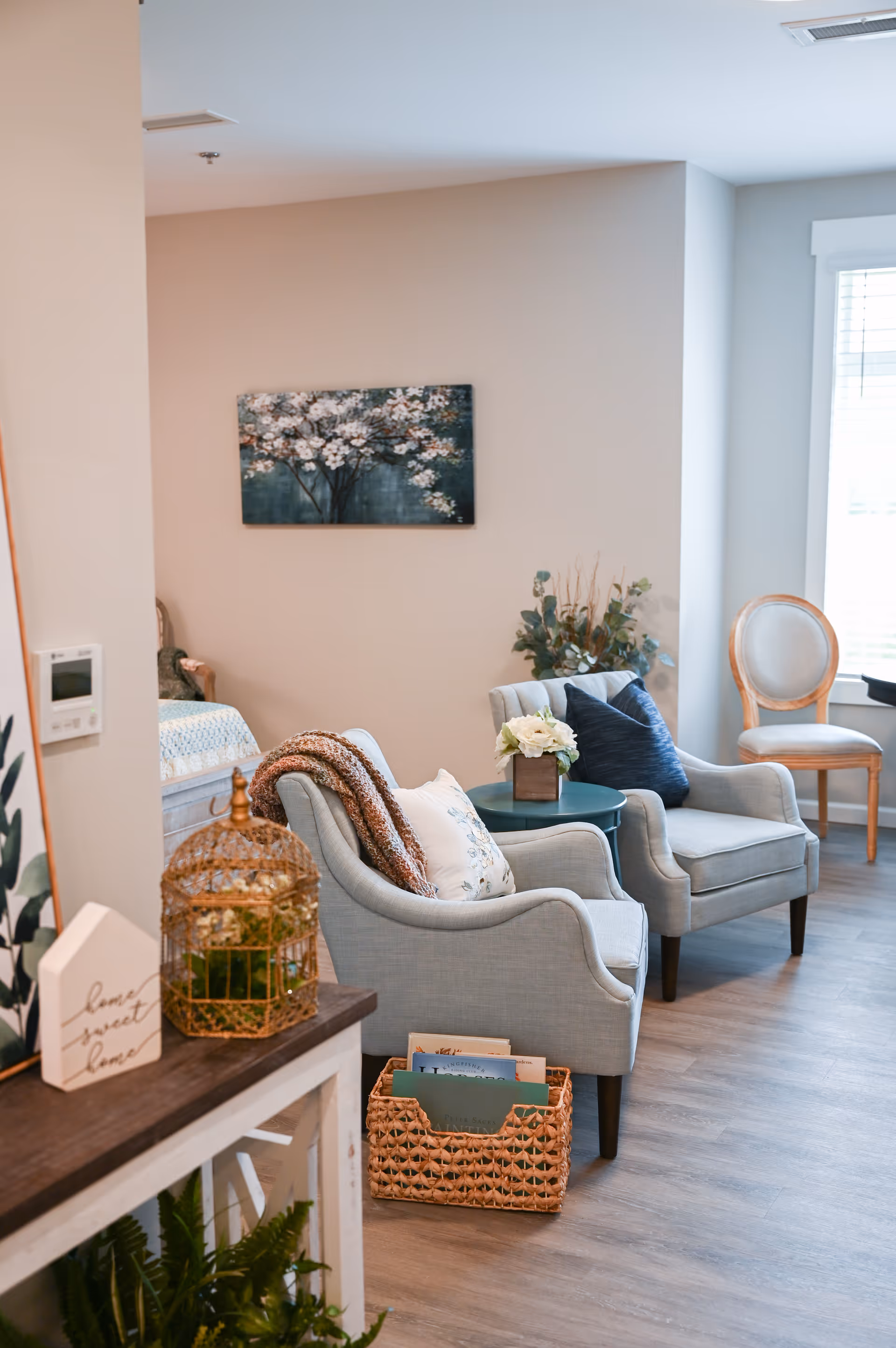 A cozy sitting area in a senior living facility with two light gray armchairs adorned with pillows and a throw blanket, a small round side table with a flower arrangement, a wicker basket with magazines or books on the floor, a decorative birdcage on a wooden console table, and a floral painting on the wall. There is also a wooden chair near a window with blinds, and the room has light-colored walls and wood flooring.