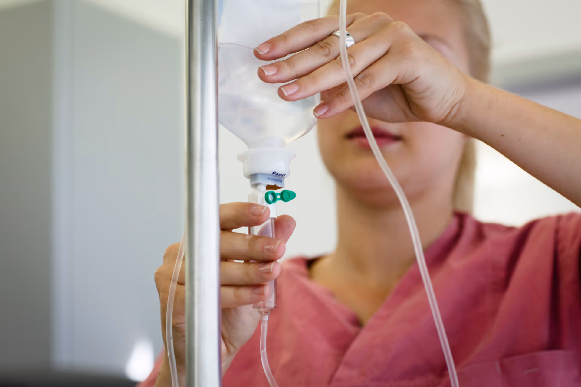 A healthcare worker in a pink uniform adjusts an intravenous (IV) drip bag, focusing on the flow regulator and tubing.