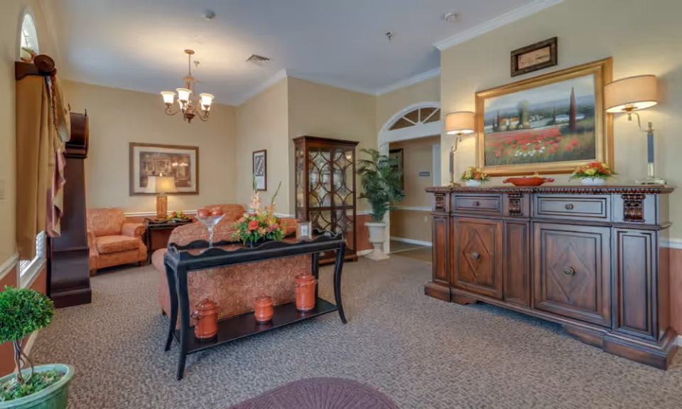 Warmly decorated senior living lounge with upholstered seating, a wooden sideboard, and decorative accents.