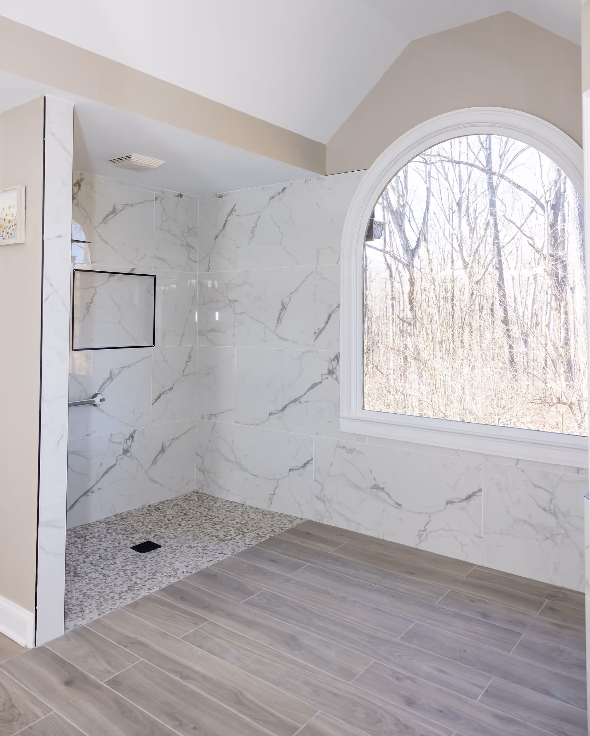 A modern bathroom shower area with marble-patterned wall tiles and a pebble stone floor. There is a large arched window letting in natural light and showing a view of leafless trees outside. The bathroom floor outside the shower is covered with wood-like tiles.