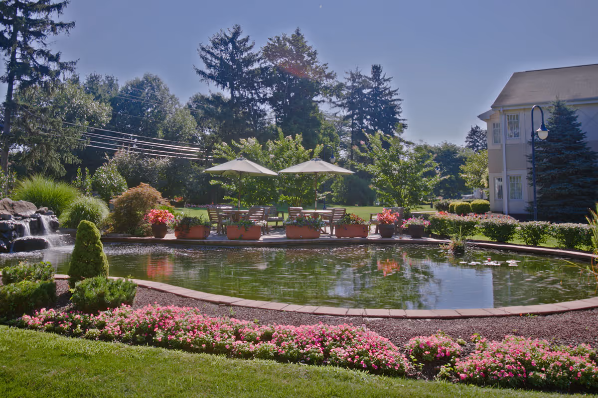 A serene outdoor garden area at Brandywine Governor's Crossing by Monarch featuring a pond with water lilies, a small waterfall, flower beds with pink flowers, potted plants, and patio tables with umbrellas surrounded by trees and greenery under a clear blue sky.
