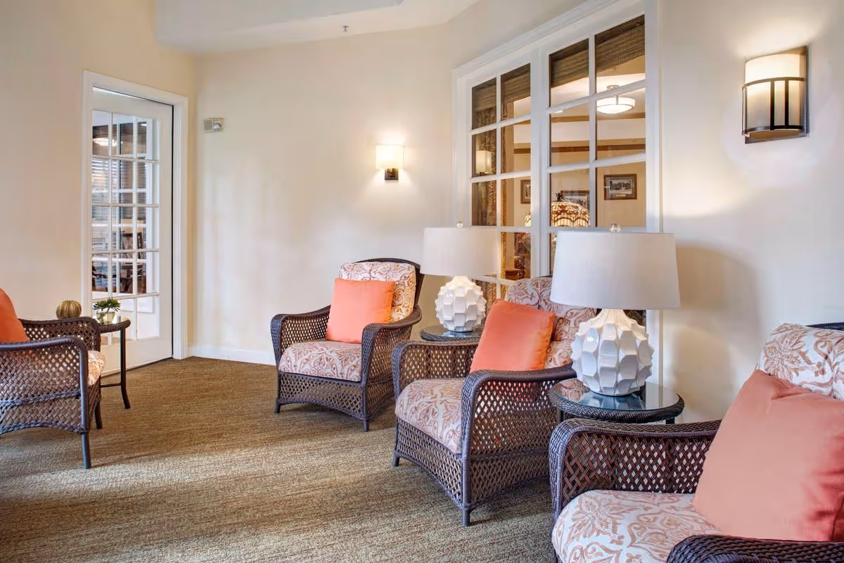 A cozy sitting area with wicker armchairs upholstered in patterned fabric and coral pillows, round side tables with lamps, and a glass-paned door/window.