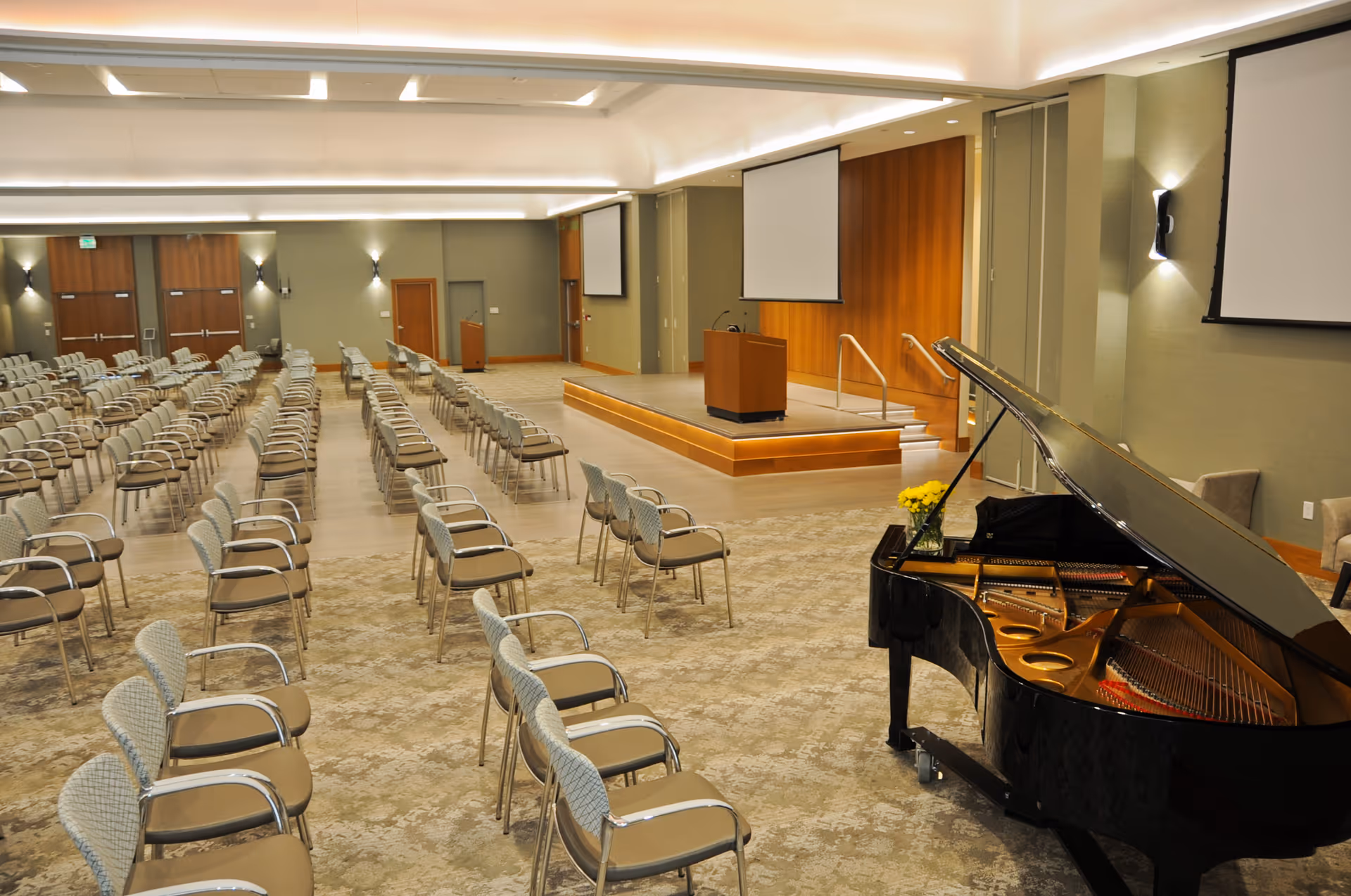 A large conference or event room with rows of chairs arranged facing a stage with a podium. The room features two large projection screens on the walls and a grand piano with a vase of yellow flowers on top in the foreground. The walls are light green with wooden accents and the floor is carpeted.