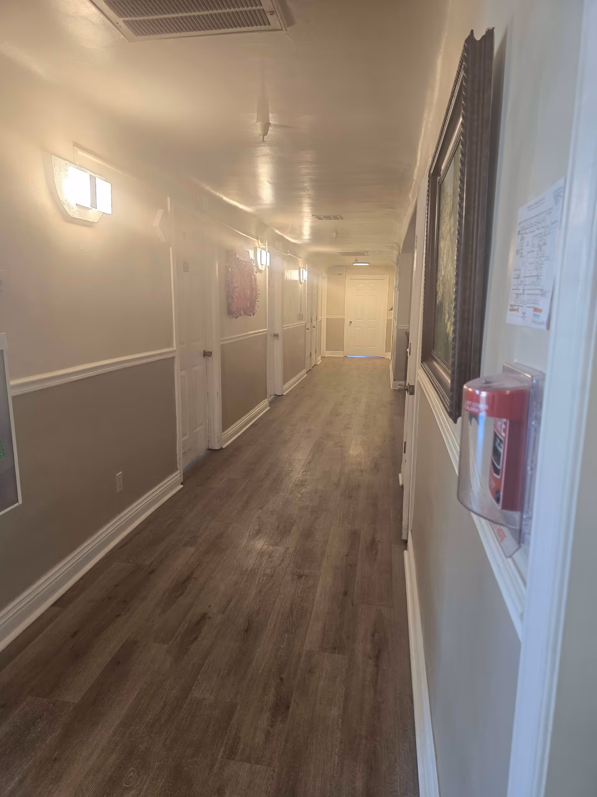 A long, narrow hallway in a senior living facility with wood-style flooring and beige walls. Several closed white doors line the left side of the hallway, and wall-mounted lights illuminate the space. On the right wall, there is a framed picture and a red fire alarm pull station with a small notice above it. The hallway extends to a closed door at the far end.