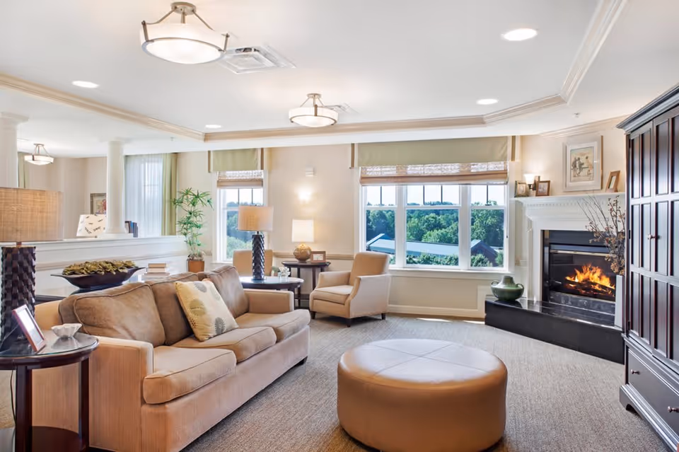 A bright and cozy living room with a beige sofa, an armchair, a round ottoman, and a fireplace with a fire burning. Large windows let in natural light and offer a view of greenery outside. The room is decorated with lamps, plants, framed pictures, and a dark wooden cabinet.