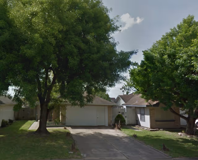 A single-story residential house with a driveway and a garage, flanked by two large leafy trees on a sunny day with a partly cloudy sky.