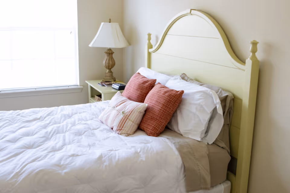 A neatly made bed with a light green wooden headboard, white bedding, and three decorative pillows in shades of red and beige. Next to the bed is a matching light green nightstand with a classic table lamp and some small items on it. A window with white blinds is visible on the left side, allowing natural light into the room.