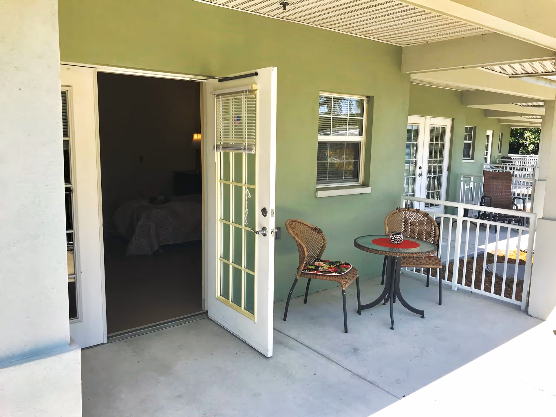 Outdoor patio area of a senior living facility with a small round table and two wicker chairs with cushions. The patio has a white railing and is adjacent to a green exterior wall with a window and an open glass door leading inside to a dimly lit bedroom.