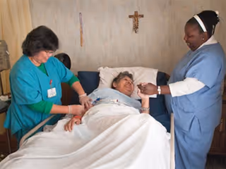 A patient lying in a hospital bed smiling while two healthcare workers, one in green scrubs and the other in blue scrubs, attend to her in a clinical room with a crucifix on the wall.
