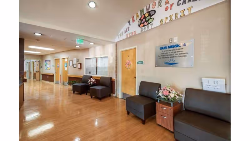 A clean and well-lit hallway in a senior living facility with wooden floors and beige walls. The hallway features several closed wooden doors, framed certificates or artwork on the walls, and seating areas with dark brown cushioned chairs and small wooden tables adorned with flower arrangements. Above one door, colorful letters spell out words related to teamwork and spirit. A mission statement plaque is mounted on the wall.