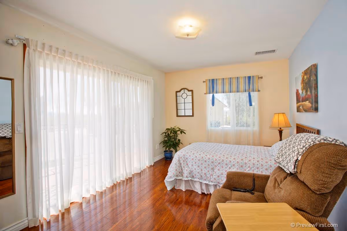 Sunny bedroom with a bed, recliner, lamp, potted plant, and floor-to-ceiling sheer curtains over sliding glass doors.