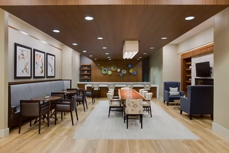 Spacious, well-lit communal dining and lounge area with a long wooden dining table, smaller round tables, upholstered chairs, and a TV nook.