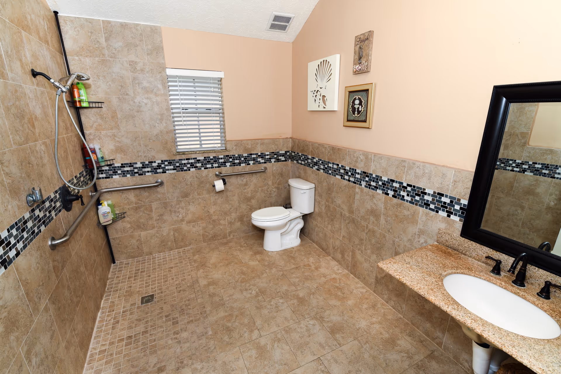 A spacious bathroom with beige tiled walls and floor, featuring a walk-in shower area with a handheld showerhead and grab bars, a white toilet, a granite countertop with a sink, and a large black-framed mirror. There is a small window with blinds and decorative wall art above the toilet.