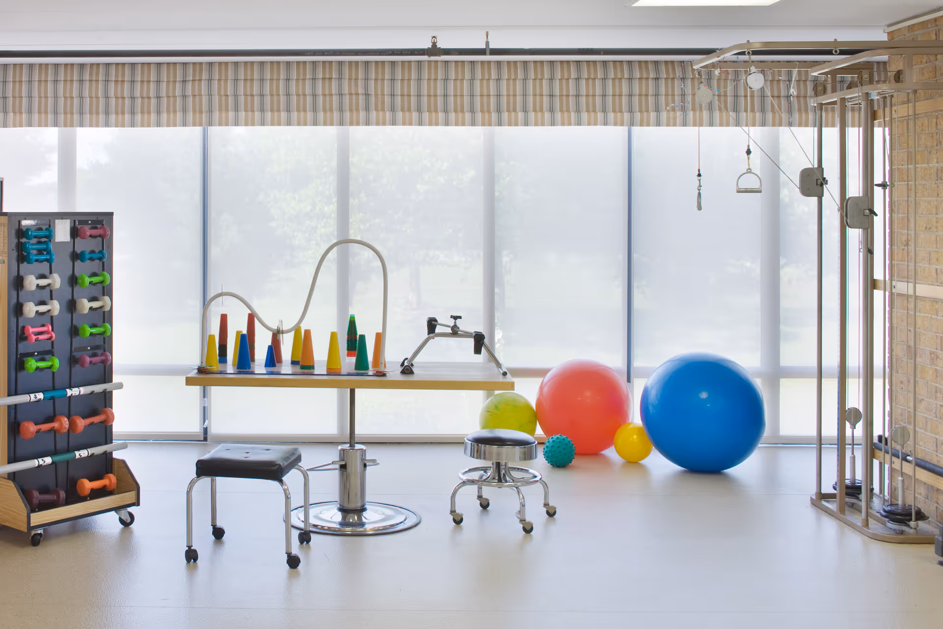 A bright physical therapy or exercise room with various equipment including colorful dumbbells on a rack, a table with cones and a hand exercise device, two stools on wheels, and several large exercise balls in red, blue, yellow, and green. The room has large windows covered with sheer blinds allowing natural light to fill the space.
