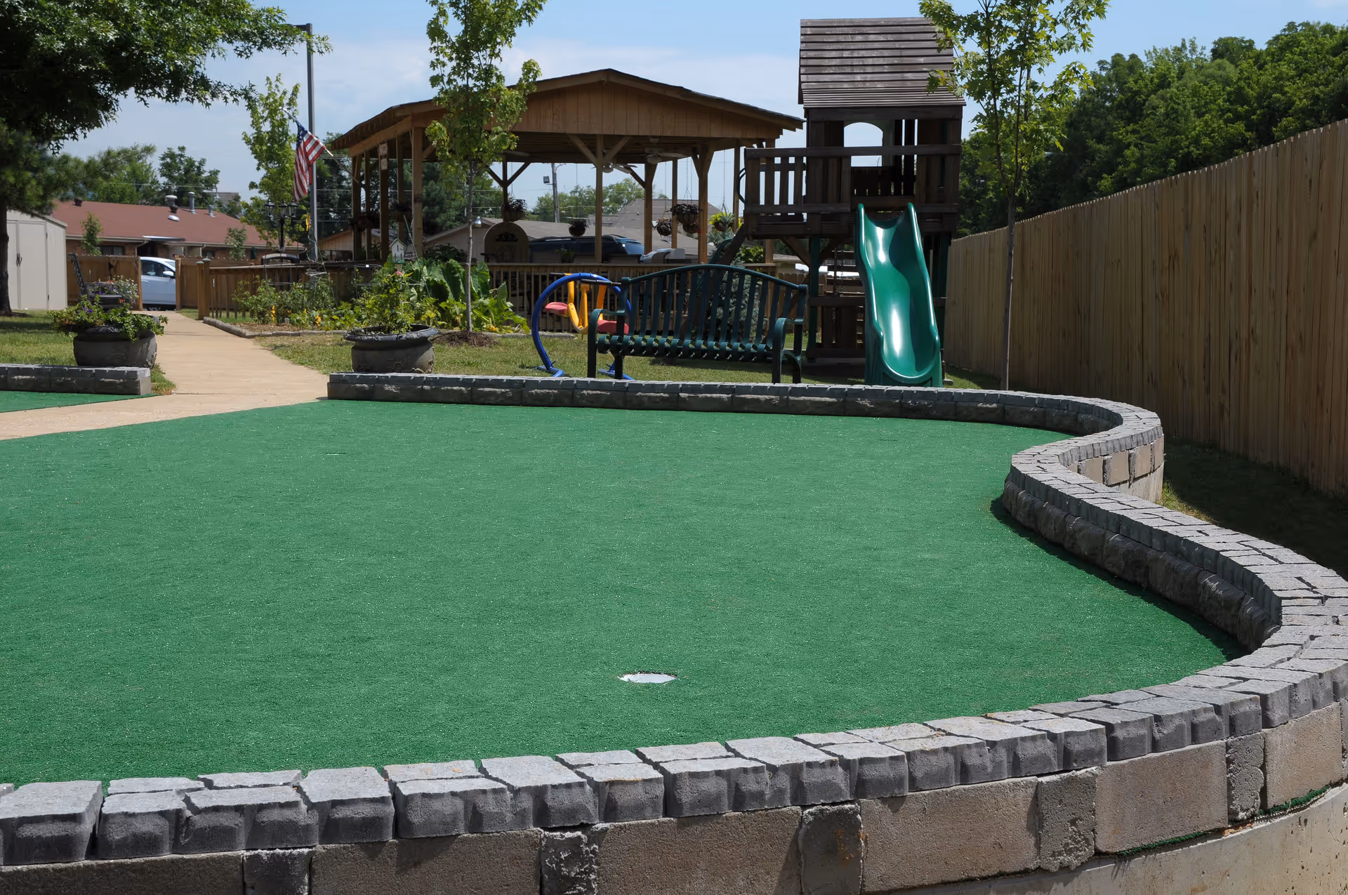 Outdoor area with a small artificial turf putting green surrounded by a low stone wall. In the background, there is a wooden playground structure with a green slide, a bench, a covered pavilion, and some trees. A wooden fence runs along the right side of the image.