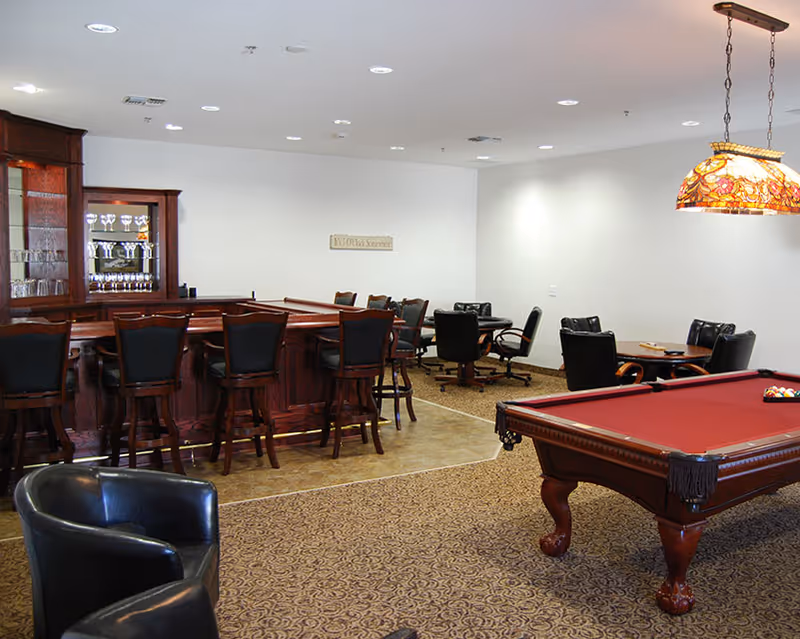 A senior living facility common area featuring a red pool table with a stained glass hanging light above it, a wooden bar with several black cushioned bar stools, and multiple seating areas with black chairs and tables. The room has a patterned carpet and a tiled floor section near the bar.