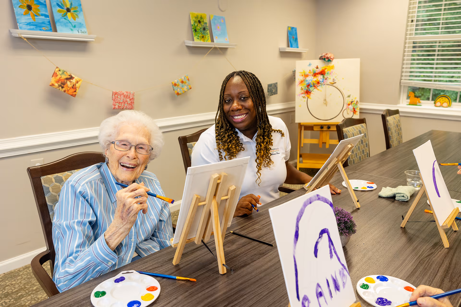 An elderly woman and a caregiver sitting at a table in a senior living facility, smiling and painting on canvases set on small easels. The room is decorated with colorful artwork on the walls and a window with blinds letting in natural light.