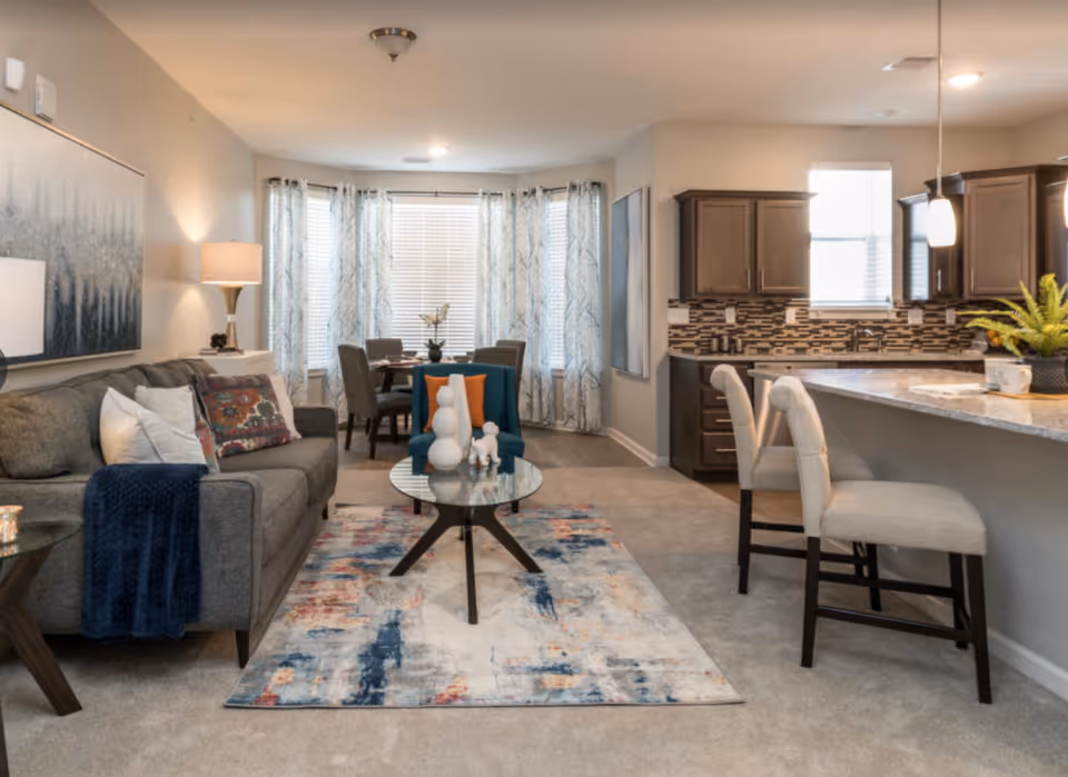 A modern open-concept living area in a senior living facility featuring a gray sofa with colorful pillows and a blue throw, a glass coffee table with decorative items, a patterned area rug, a blue armchair with an orange pillow, a dining table with four chairs near large windows with sheer curtains, and a kitchen area with a breakfast bar and two upholstered bar stools.