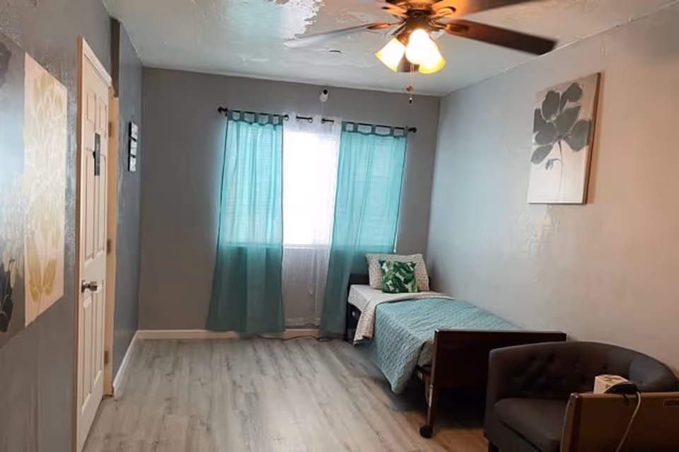 A small bedroom with gray walls and light wood flooring. There is a single bed with a teal quilt and decorative pillows near a window with teal curtains. A ceiling fan with lights is on the ceiling. A gray armchair is positioned near the bed, and there are two pieces of wall art featuring floral designs.