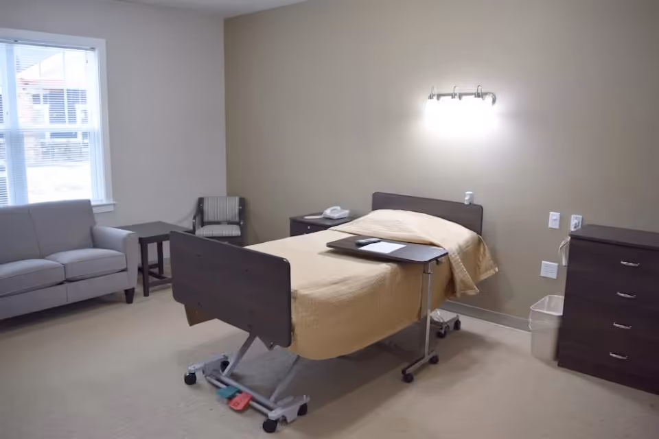 A senior living facility bedroom with a single hospital-style bed covered with a beige blanket, a small overbed table with a remote and paper, a gray sofa, a striped armchair, a side table, a dark wooden dresser, and a window letting in natural light.
