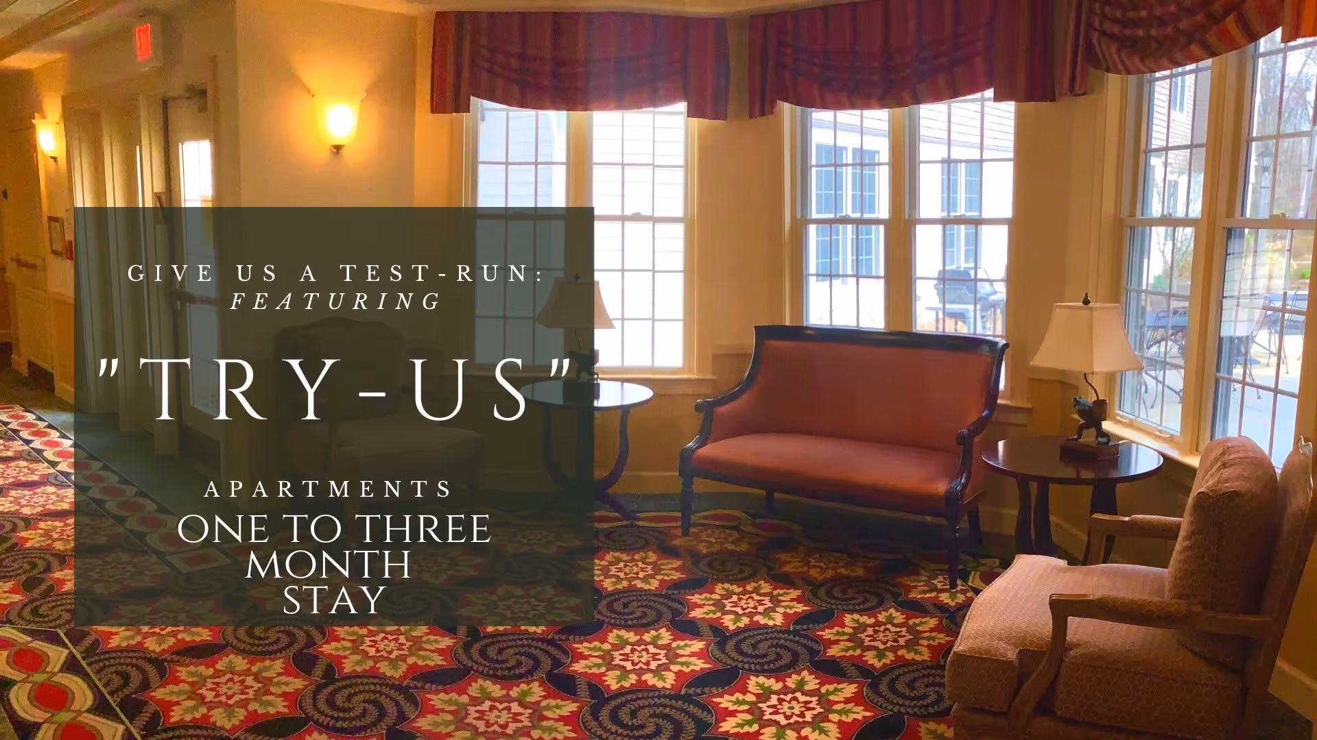 A cozy common sitting area with upholstered chairs, a loveseat, side tables and large windows over a patterned carpet.