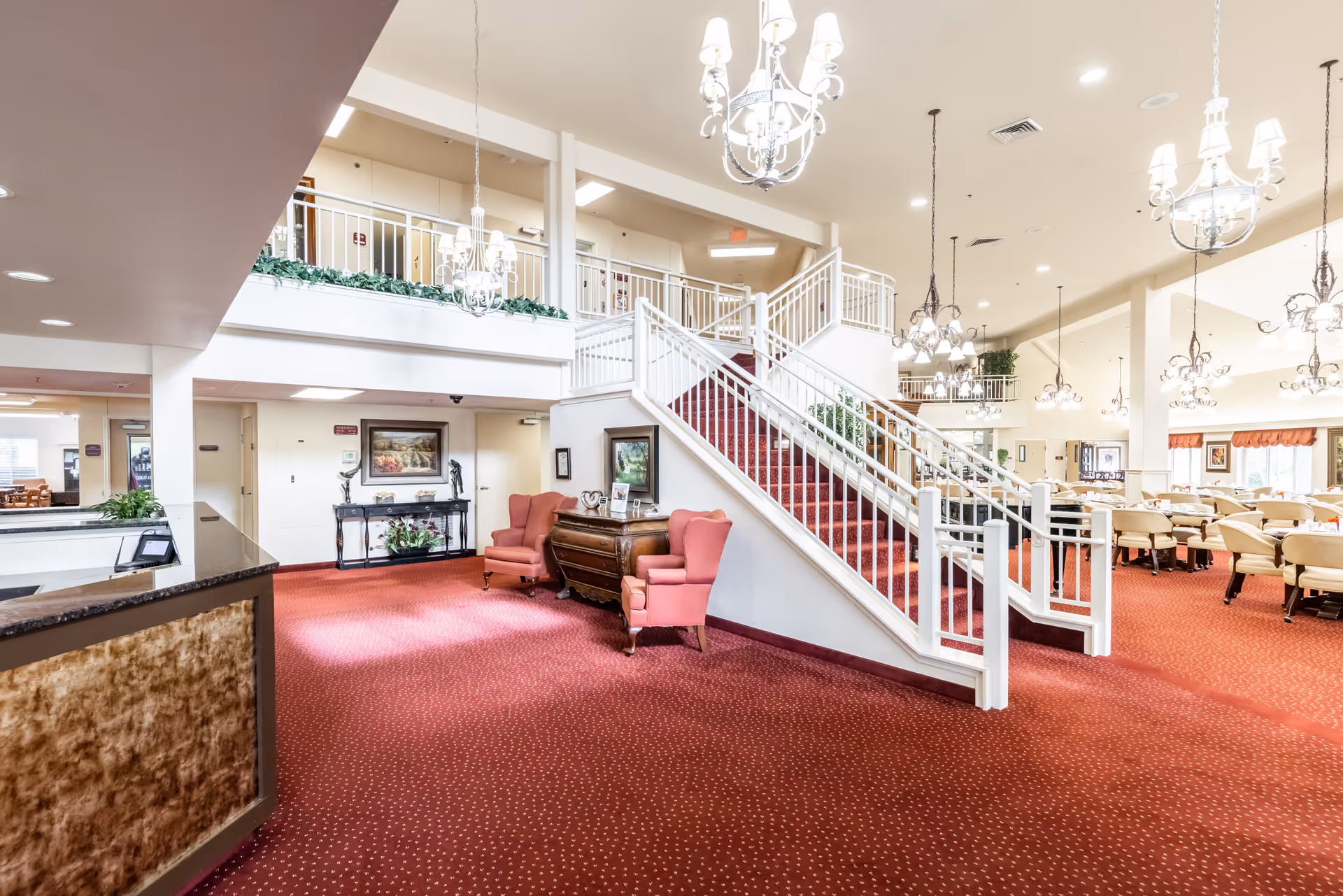 Bright and spacious senior living facility interior with red carpet flooring, a white staircase with railings leading to an upper level, multiple chandeliers hanging from the ceiling, two pink armchairs and a wooden chest of drawers near the staircase, and a dining area with tables and chairs in the background.