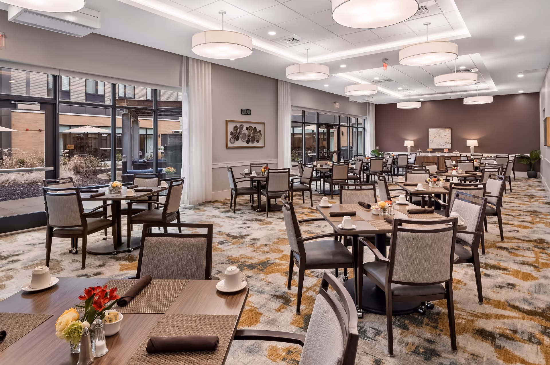 A spacious dining room with multiple tables and chairs arranged neatly. Each table is set with placemats, napkins, cups, and small flower arrangements. Large windows on one side let in natural light and offer a view of an outdoor patio area. The ceiling features modern circular light fixtures, and the walls are decorated with framed artwork. The room has a patterned carpet and a calm, neutral color palette.