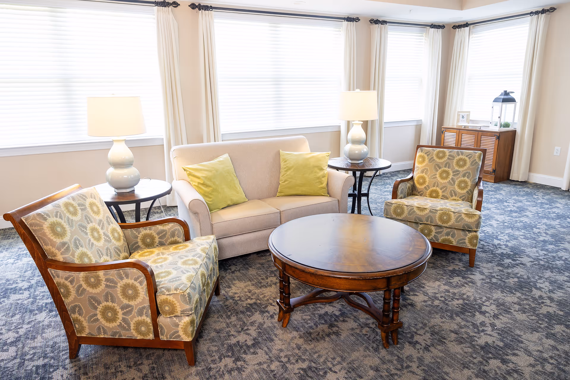 A bright and cozy living room area with a beige loveseat adorned with two green pillows, two floral-patterned armchairs with wooden frames, a round wooden coffee table in the center, two side tables each holding a white lamp, large windows with white blinds and cream curtains, and a wooden cabinet in the corner.
