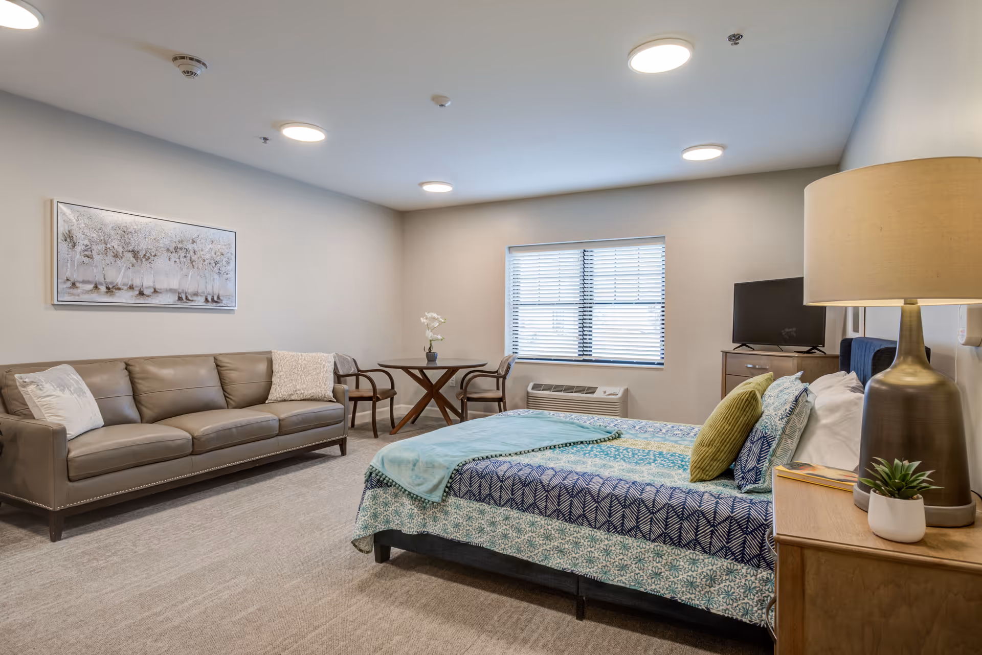 A furnished bedroom with a bed, patterned bedding, a sofa, small table and chairs, TV, and lamp in a senior living facility.