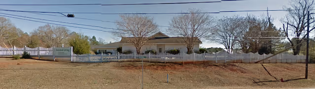 Single-story building set behind a white picket fence with trees, a lawn, and a sign on the left.