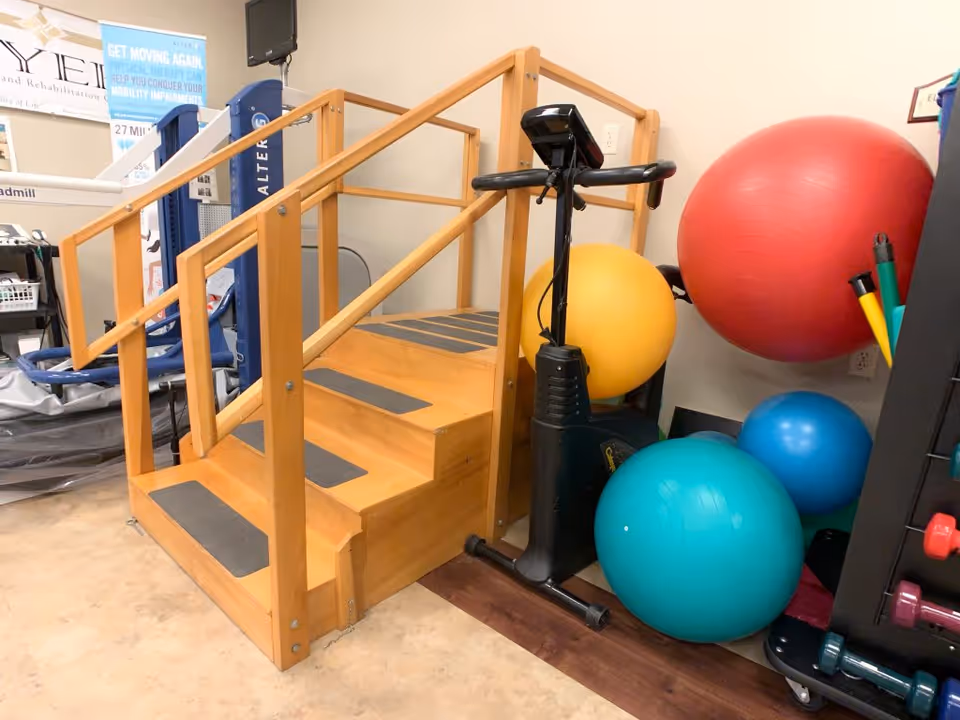 A small indoor physical therapy area featuring a wooden staircase with handrails, several large colorful exercise balls, a stationary exercise bike, and various exercise equipment including dumbbells and resistance bands.