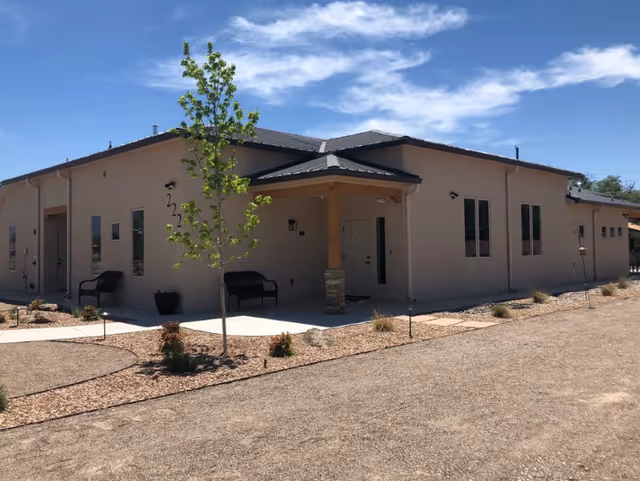 Single-story beige senior living building with a covered entrance, benches, a young tree, and xeriscaped landscaping under a blue sky.