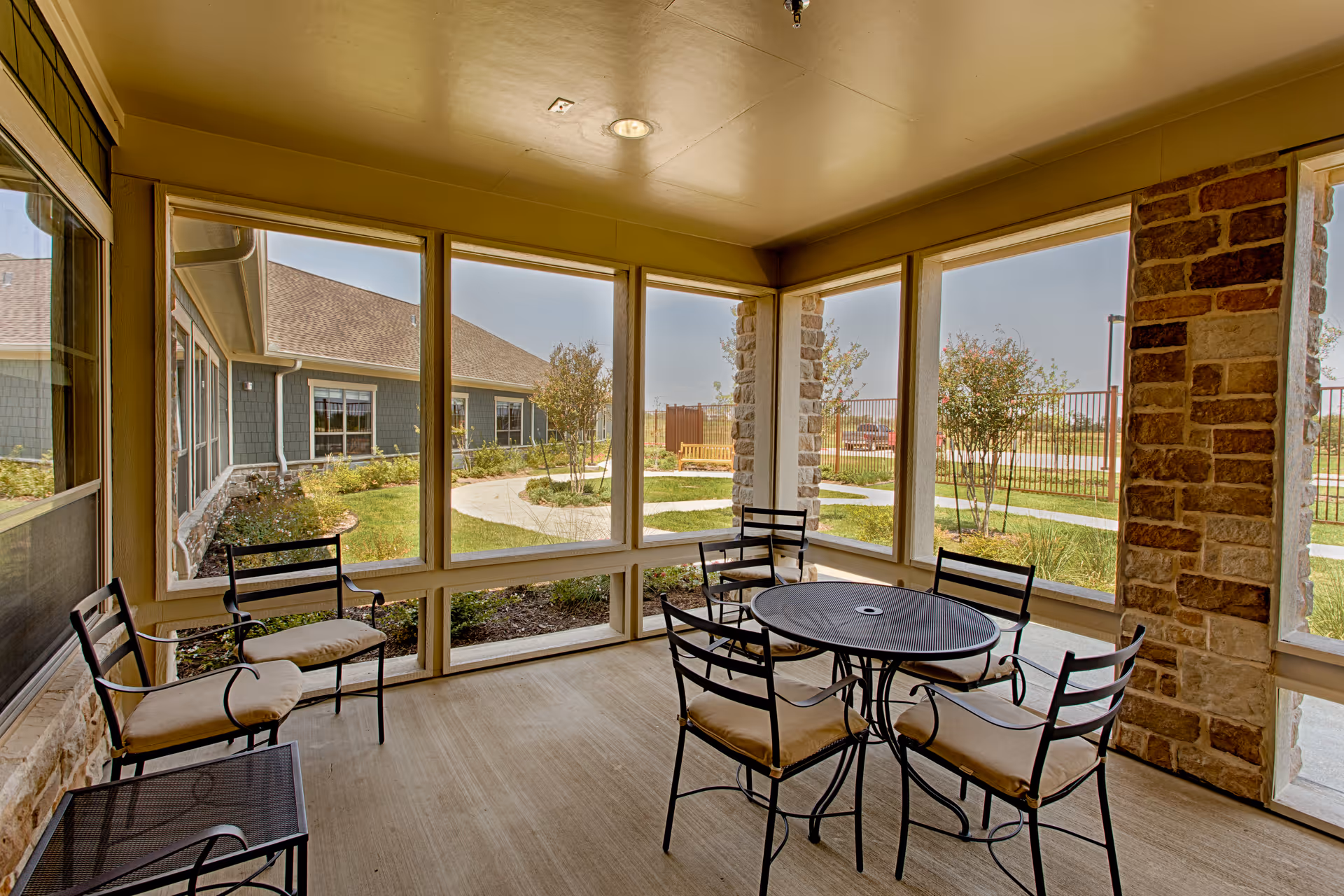 A screened-in porch area with a round metal table and six cushioned metal chairs. The porch overlooks a landscaped garden with a pathway, small trees, and a bench. The building exterior features stone pillars and siding with windows.