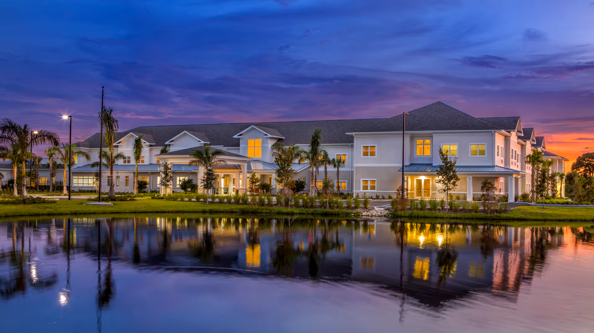 A large, two-story senior living facility building at dusk with lights glowing from the windows, surrounded by palm trees and landscaping, reflected in a calm pond in the foreground under a colorful purple and orange sky.