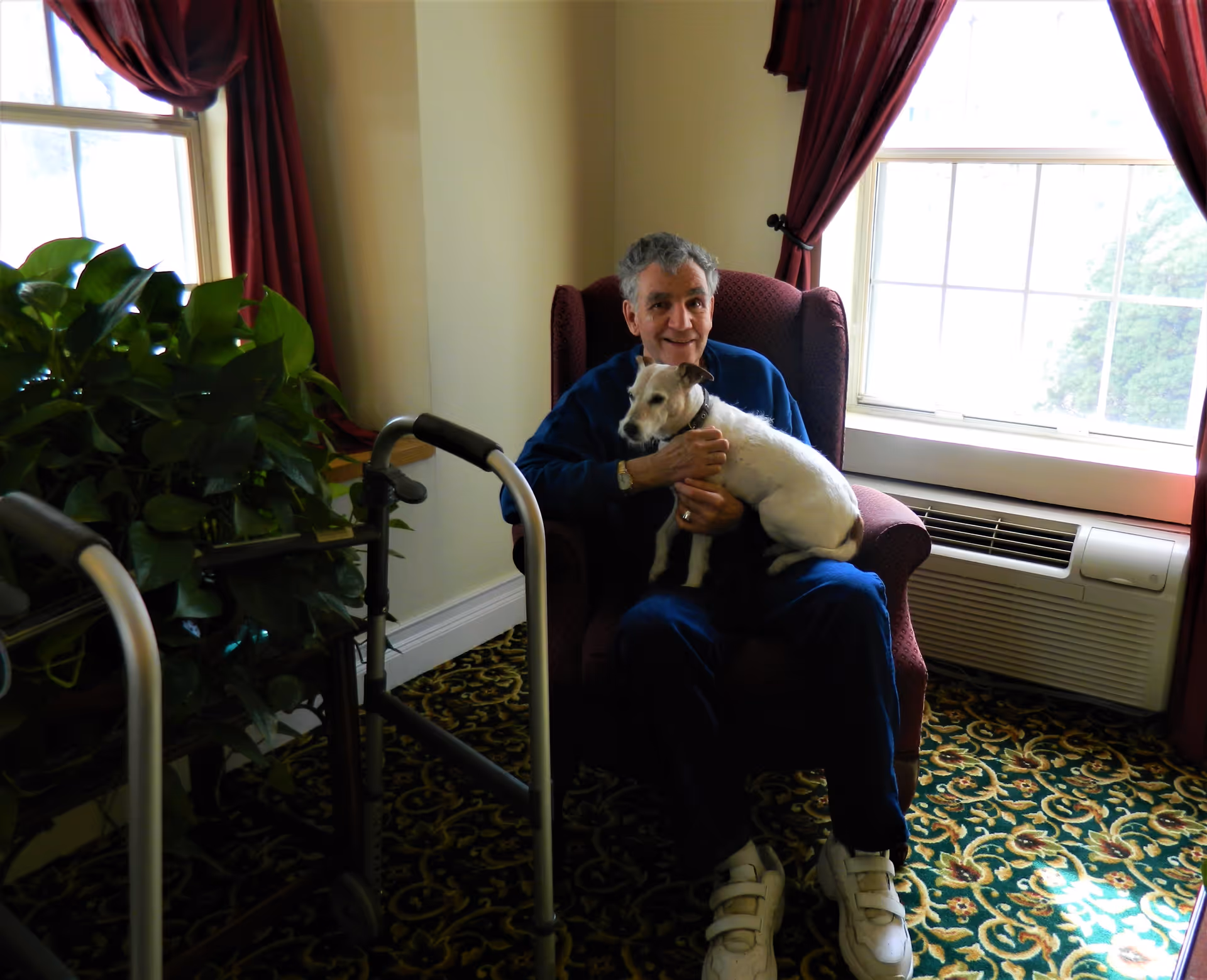 An elderly man sitting in a maroon armchair near a window with red curtains, holding a small white and brown dog on his lap. A walker and a green leafy plant are visible to the left, and an air conditioning unit is below the window.